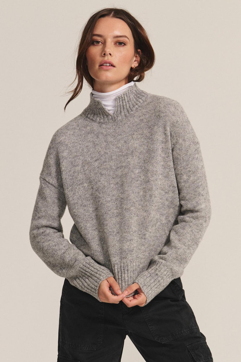 A woman stands against a plain background wearing the BRIELLE SWEATER by Velvet by Graham & Spencer over a white turtleneck and black pants, holding the sweater’s hem.