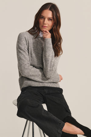A woman with long brown hair wears the BRIELLE SWEATER by Velvet by Graham & Spencer and black pants while sitting on a stool, resting her chin on her hand against a plain background.