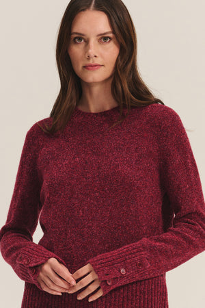 A woman wears the MAISIE SWEATER by Velvet by Graham & Spencer, featuring long sleeves, textured burgundy fabric, a crew neckline, and cuff button details, standing against a plain background.
