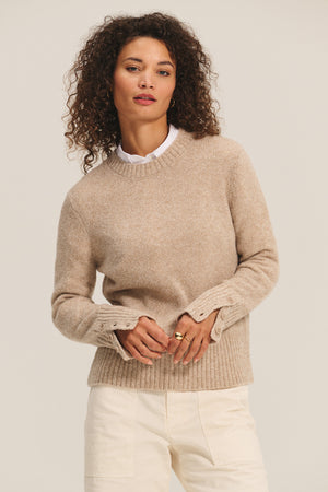 A woman with curly hair models the MAISIE SWEATER by Velvet by Graham & Spencer—a beige wool blend crewneck—layered over a white collared shirt and light pants, standing against a plain background.