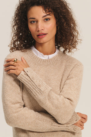 A woman with curly hair wears the Velvet by Graham & Spencer MAISIE SWEATER, a beige wool blend with buttoned cuffs, layered over a white collared shirt, and looks at the camera with a neutral expression.