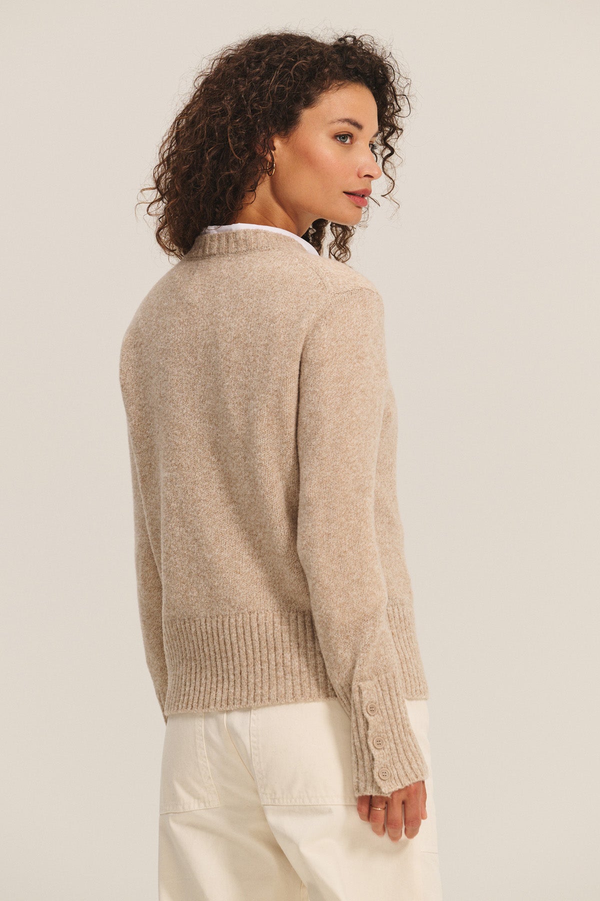 A woman with curly hair wears the MAISIE SWEATER by Velvet by Graham & Spencer—a beige wool blend with a crew neckline—and white pants, standing slightly turned from the camera against a plain background.
