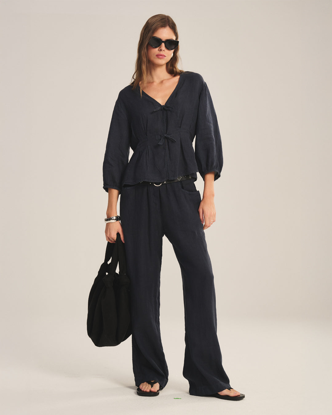 A woman in sunglasses, the Velvet by Graham & Spencer MARTHA LINEN TOP with pleated details, matching wide-leg pants, sandals, and a large black bag stands against a neutral background.-41088977895617