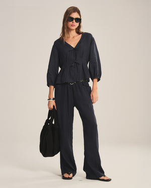 A woman in sunglasses, the Velvet by Graham & Spencer MARTHA LINEN TOP with pleated details, matching wide-leg pants, sandals, and a large black bag stands against a neutral background.