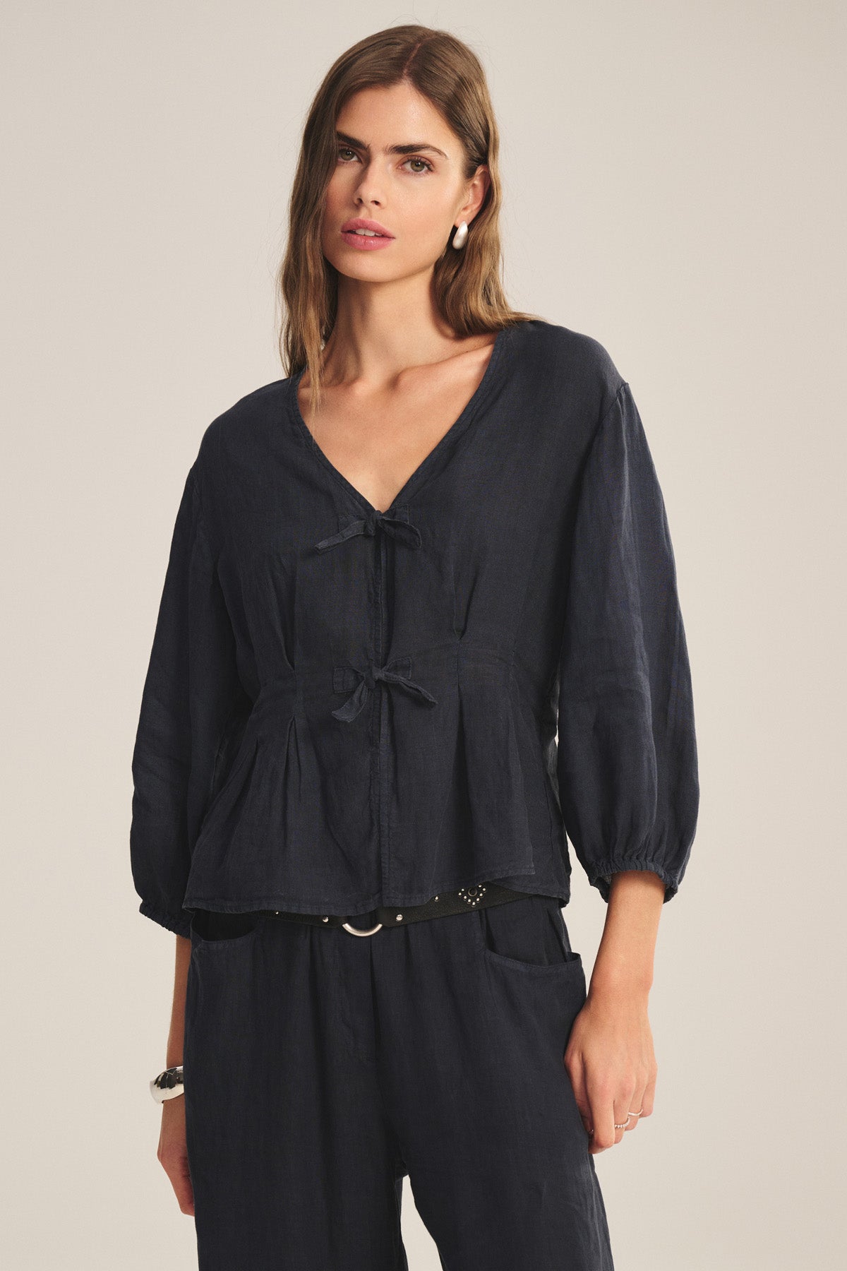 A woman wears the MARTHA LINEN TOP by Velvet by Graham & Spencer— a loose-fitting, long-sleeve blouse with puff sleeves and a front tie—paired with matching dark pants, standing against a neutral background.-41089020461249