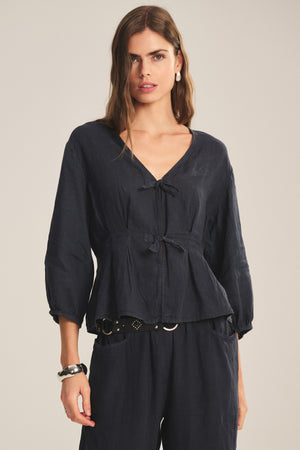 A woman models the MARTHA LINEN TOP by Velvet by Graham & Spencer, featuring puffed sleeves and tie closures, styled with matching dark pants against a plain background.