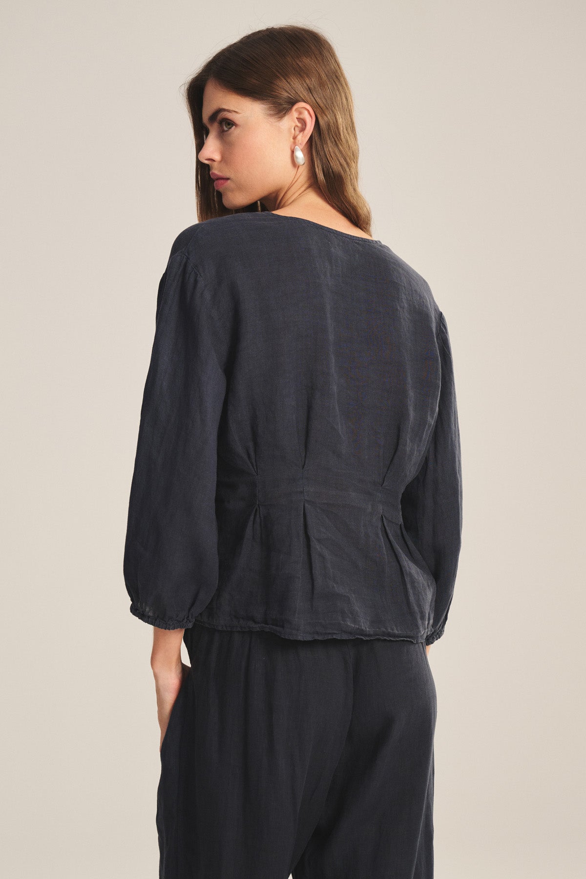 A woman with long brown hair is seen from the back wearing the Velvet by Graham & Spencer MARTHA LINEN TOP, a loose-fitting blouse with puffed sleeves, paired with matching pants against a plain light background.