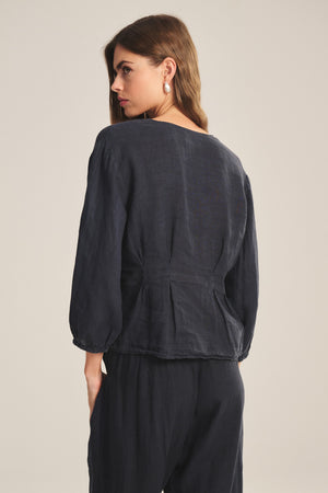 A woman with long brown hair is seen from the back wearing the Velvet by Graham & Spencer MARTHA LINEN TOP, a loose-fitting blouse with puffed sleeves, paired with matching pants against a plain light background.