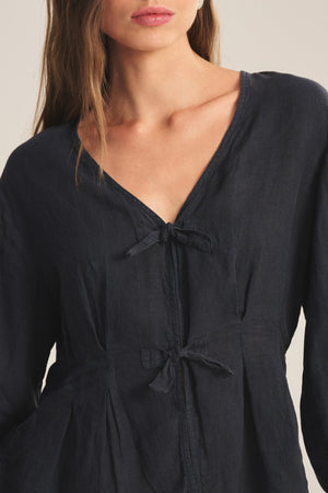 A woman in the MARTHA LINEN TOP by Velvet by Graham & Spencer—a dark, breathable linen top with two front bow ties, pleated details, and a V-neckline. Her face is partially visible.