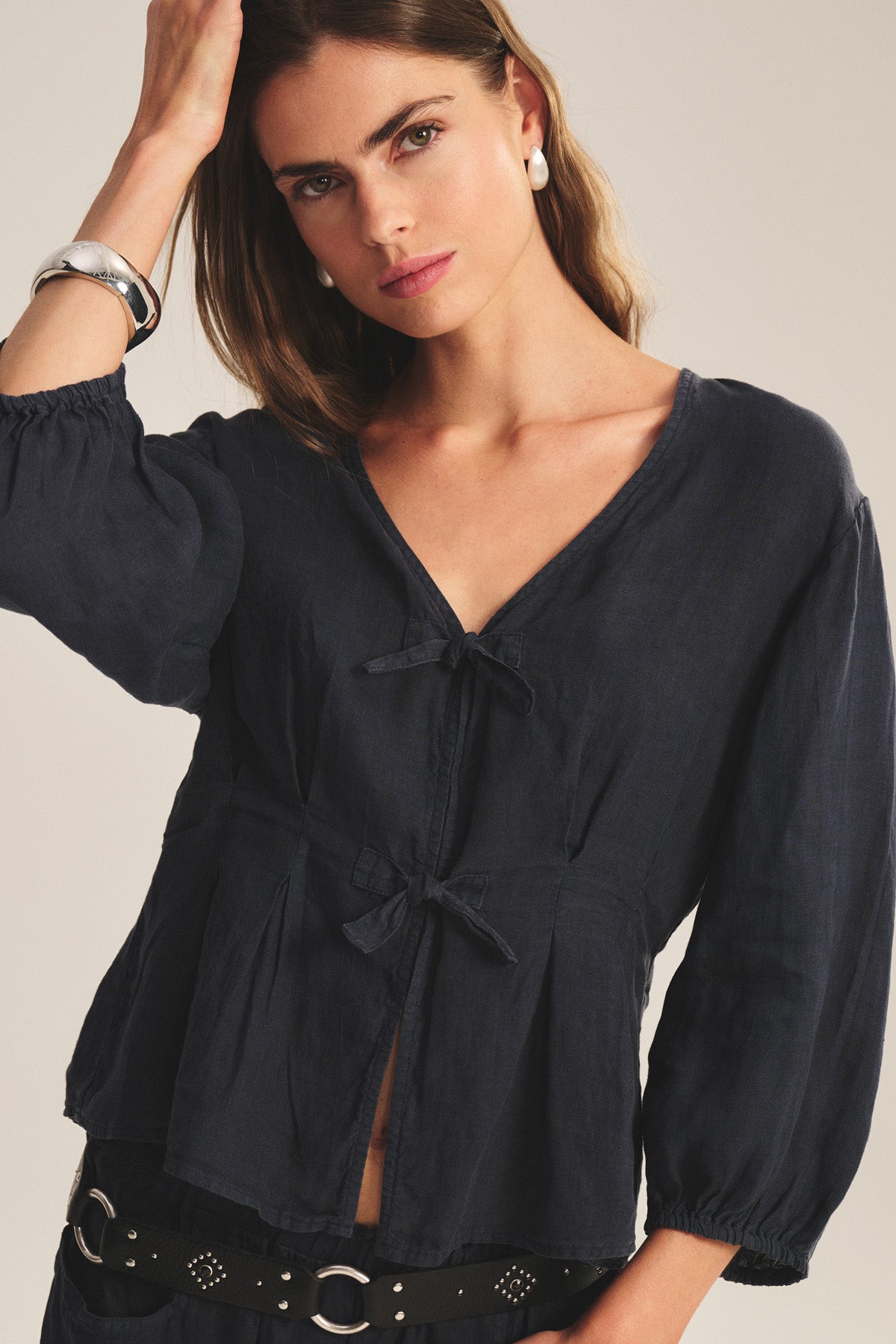 A woman wears the MARTHA LINEN TOP by Velvet by Graham & Spencer—a navy, long-sleeve, pleated V-neck blouse with a tied front—paired with black pants, a studded belt, pearl earrings, and a silver bracelet against a plain background.