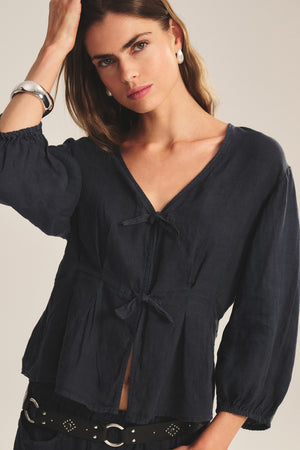 A woman wears the MARTHA LINEN TOP by Velvet by Graham & Spencer—a navy, long-sleeve, pleated V-neck blouse with a tied front—paired with black pants, a studded belt, pearl earrings, and a silver bracelet against a plain background.