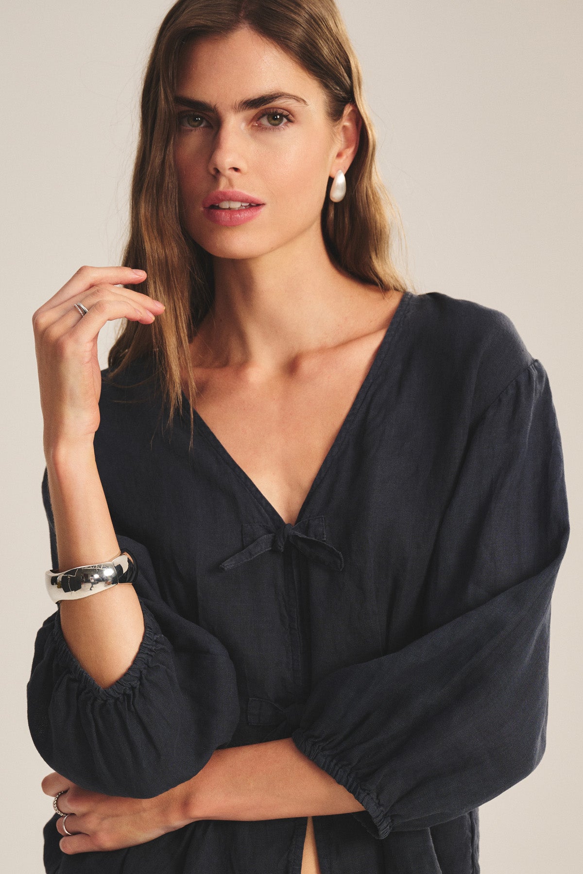 A woman with long brown hair models the MARTHA LINEN TOP by Velvet by Graham & Spencer, featuring puffed sleeves, styled with large pearl earrings and a chunky silver bracelet against a neutral background.
