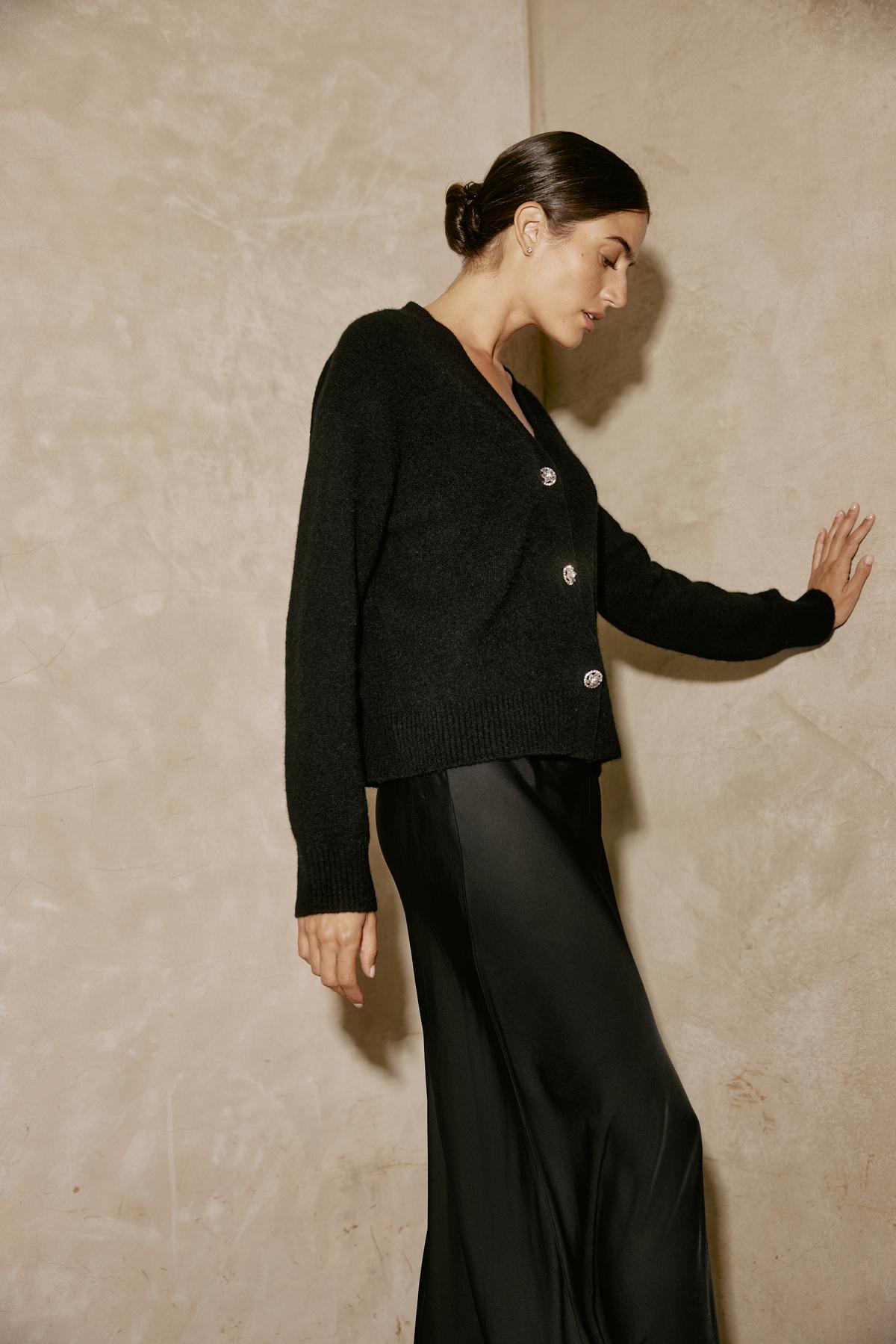   A person wearing the MARTI ALPACA EMBELLISHED CARDIGAN by Velvet by Graham & Spencer and a satin skirt leans against a textured wall. 