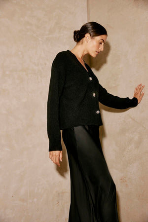 A person wearing the MARTI ALPACA EMBELLISHED CARDIGAN by Velvet by Graham & Spencer and a satin skirt leans against a textured wall.