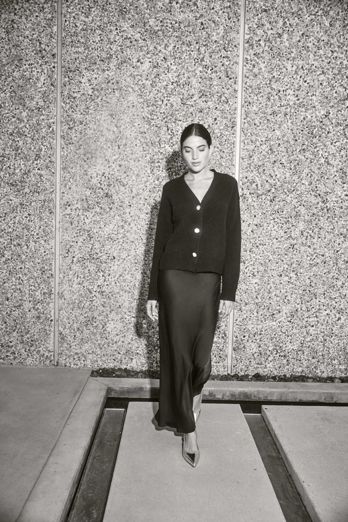   A person wearing the MARTI ALPACA EMBELLISHED CARDIGAN by Velvet by Graham & Spencer and a long skirt walks confidently beside a textured wall in a minimalist setting. 