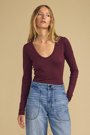 A woman wearing the MATILDA TEE by Velvet by Graham & Spencer—a long-sleeve, scoop neck maroon top—paired with high-waisted blue jeans stands against a beige background, showcasing this perfect layering piece for any season.