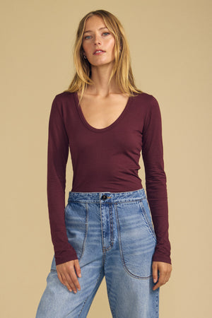 A woman stands against a beige background, wearing the Velvet by Graham & Spencer MATILDA TEE—a maroon scoop neck long sleeve top—paired with high-waisted blue jeans.