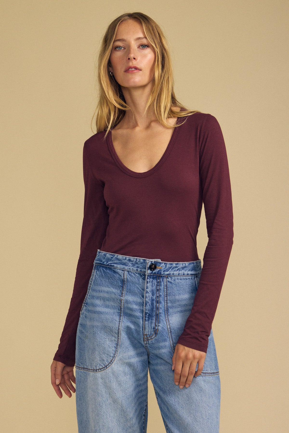 A woman wearing the MATILDA TEE by Velvet by Graham & Spencer—a maroon scoop neck long sleeve top—paired with high-waisted blue jeans stands in front of a plain beige background, showcasing this essential layering piece for any wardrobe.
