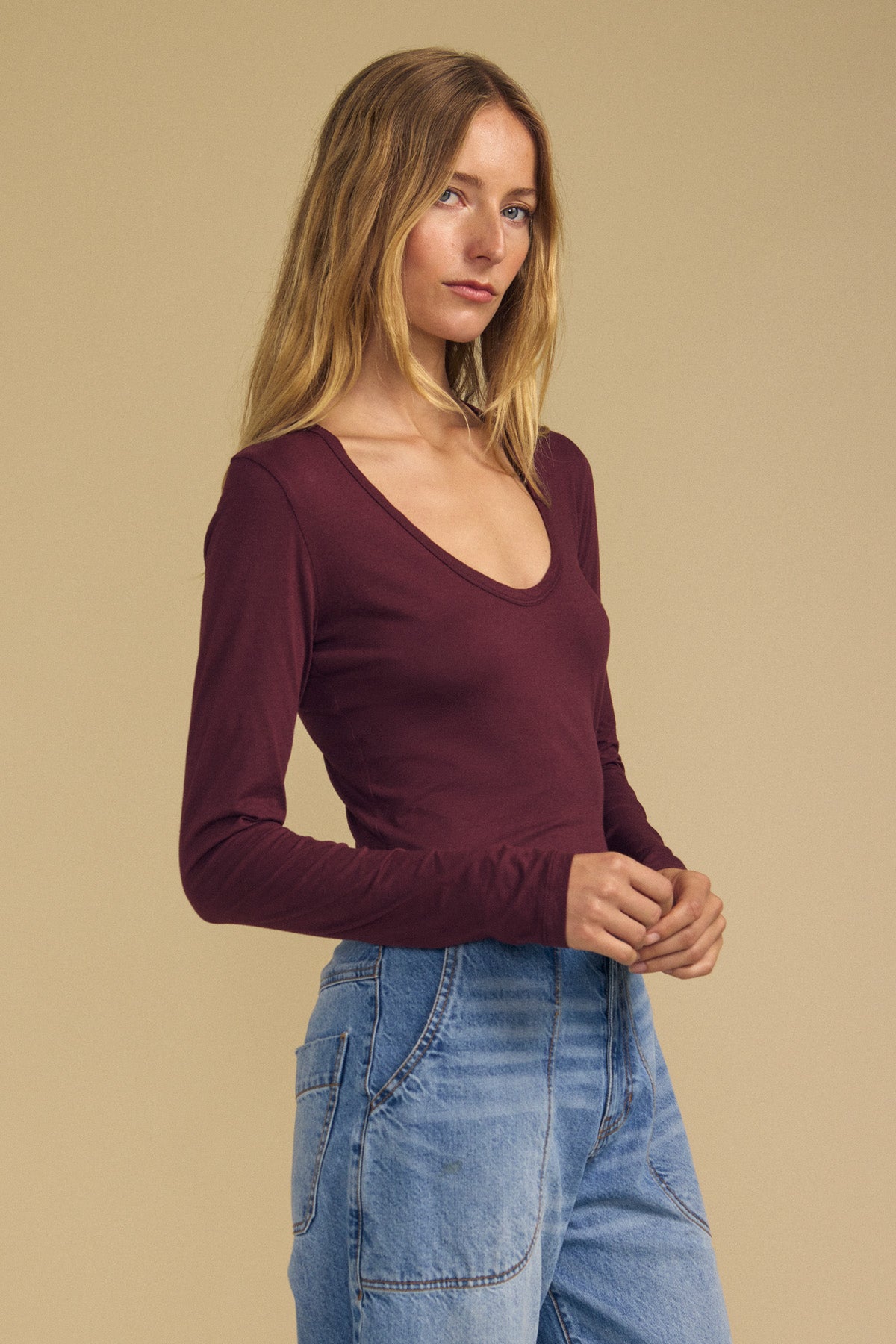 A woman with long blonde hair wears the Velvet by Graham & Spencer MATILDA TEE—a burgundy scoop neck, long sleeve top—paired with blue jeans and standing against a plain beige background. This versatile tee is the perfect layering piece for any wardrobe.