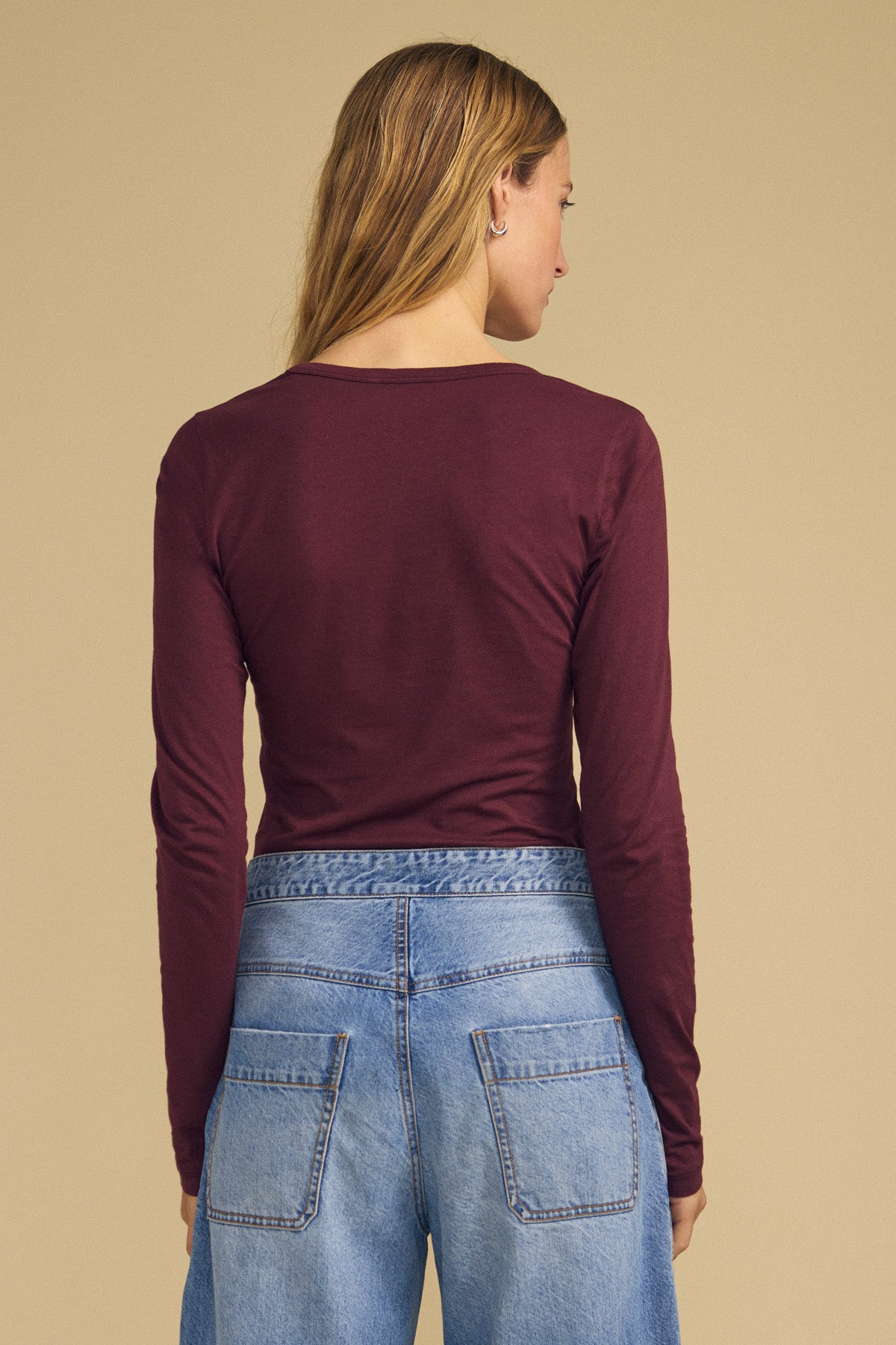 Wearing the MATILDA TEE by Velvet by Graham & Spencer in maroon, a person with long hair is shown from the back layering this long sleeve top with blue jeans against a plain beige background.