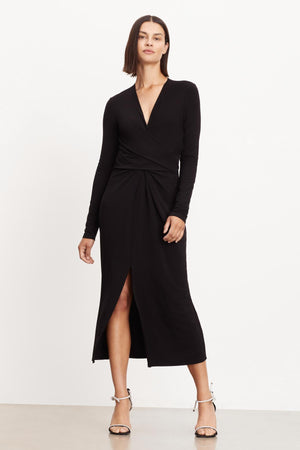 A model wearing an ELIANA WRAP MIDI DRESS by Velvet by Graham & Spencer, featuring a black matte jersey fabric, a thigh-high slit, and a v-neckline.
