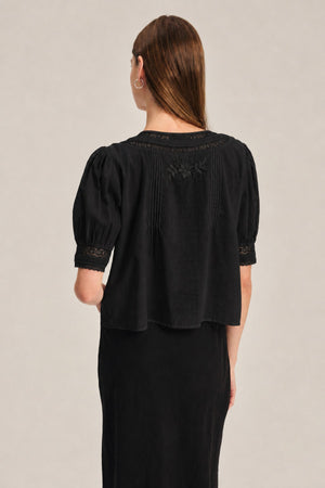 A person with straight brown hair wears the MAVIS TOP by Velvet by Graham & Spencer—a feminine sheer black short-sleeve top with lace and delicate embroidery—shown from the back.