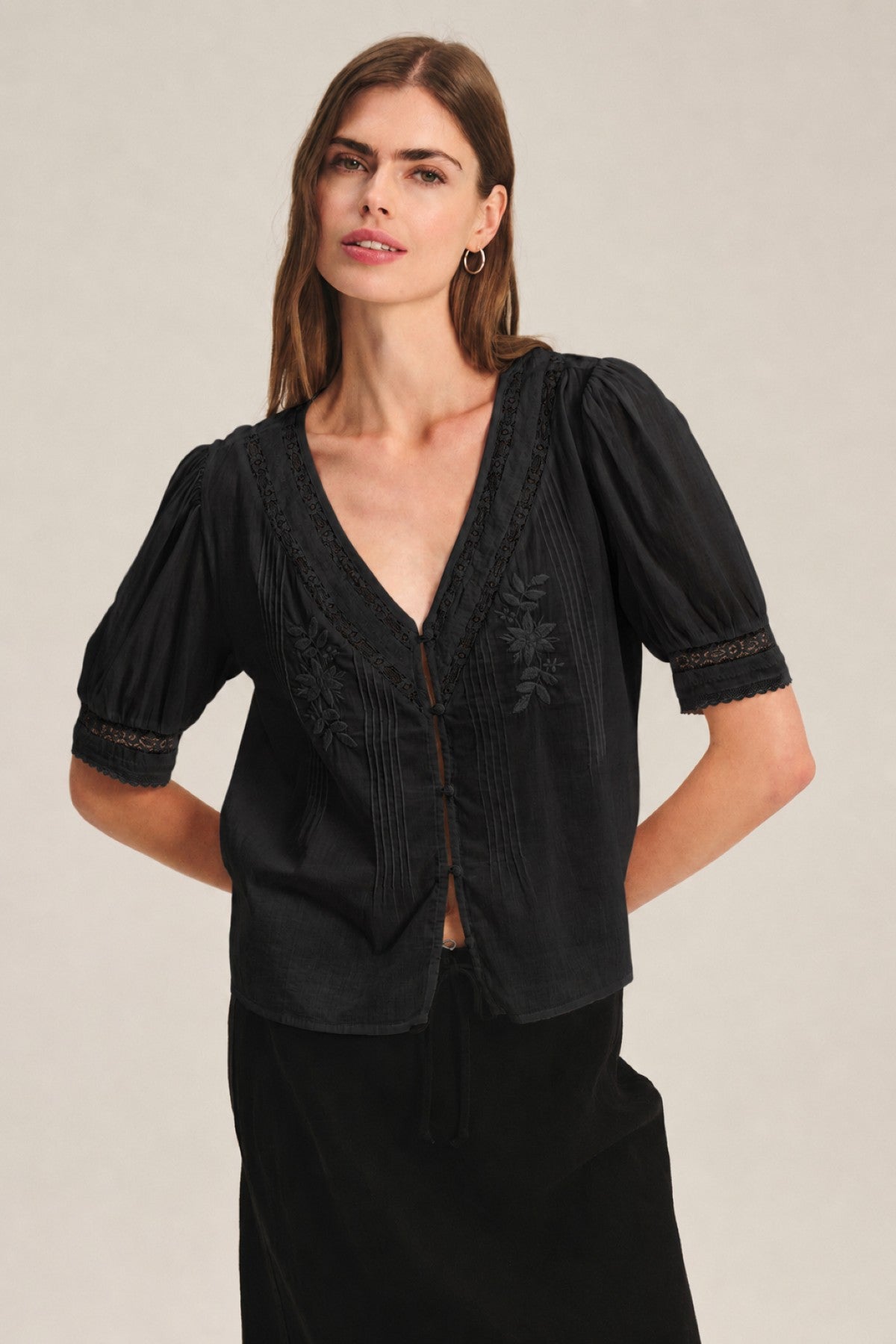 A woman wears the MAVIS TOP by Velvet by Graham & Spencer—a black short-sleeve blouse with lace trim and delicate embroidery—paired with a black skirt, standing against a plain background.