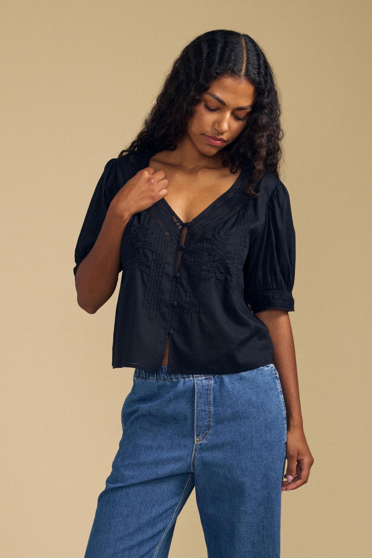 A woman with long curly hair wears the MAVIS TOP by Velvet by Graham & Spencer—a feminine lace blouse with short sleeves—paired with blue jeans, standing against a beige background and looking downward.-41802945134785