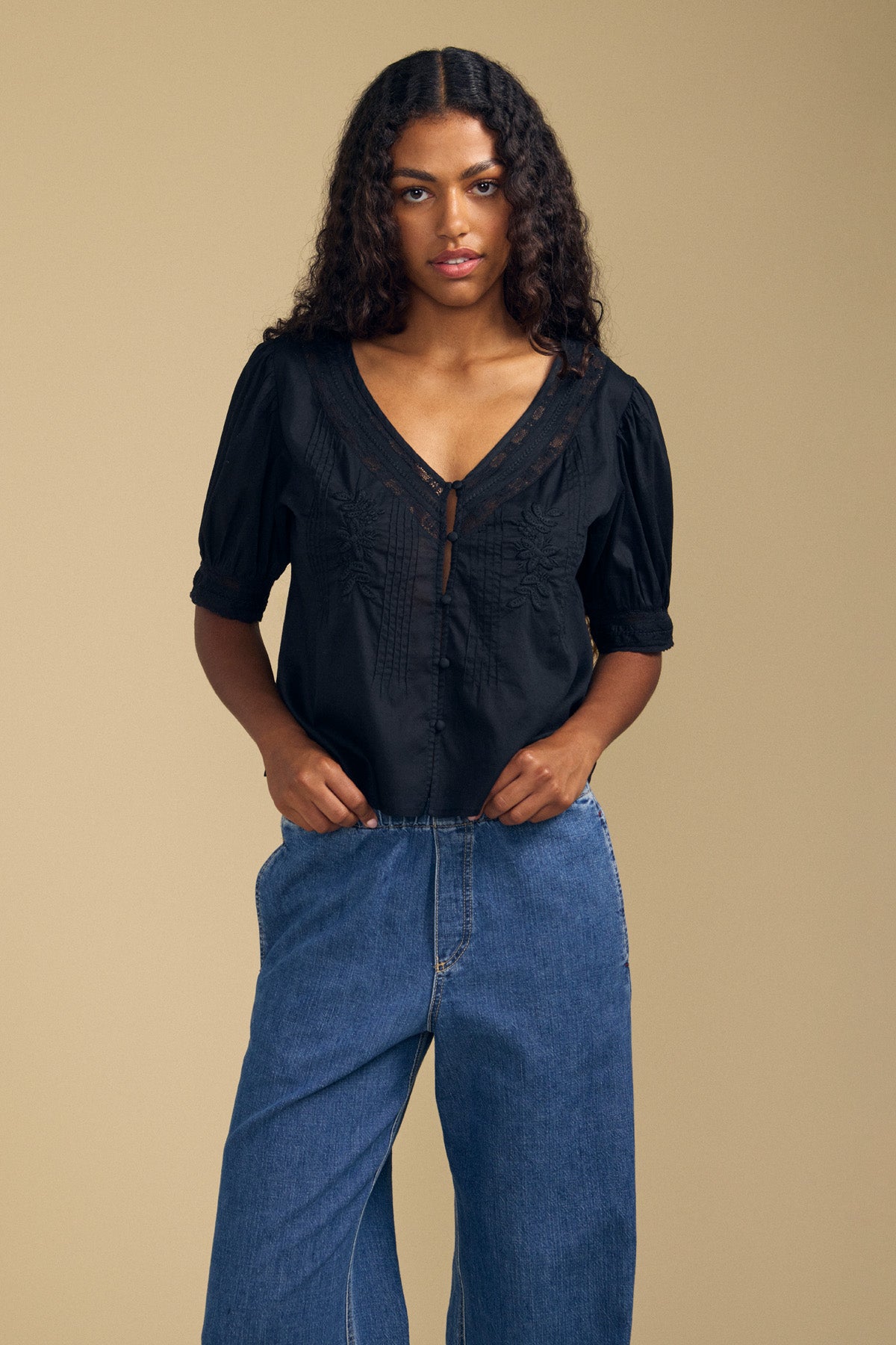 A woman with long curly hair wears the MAVIS TOP by Velvet by Graham & Spencer—a feminine short-sleeved lace blouse—paired with blue jeans, standing against a plain beige background.-41802938056897