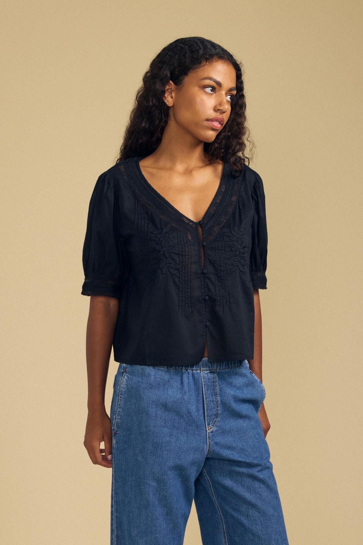 A woman wears the MAVIS TOP by Velvet by Graham & Spencer—a black blouse adorned with delicate floral appliqué and embroidery—paired with blue jeans, standing against a plain beige background.-41802945167553