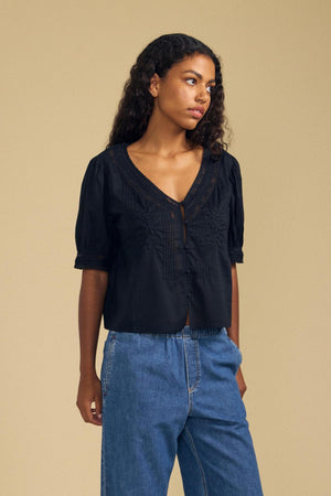 A woman wears the MAVIS TOP by Velvet by Graham & Spencer—a black blouse adorned with delicate floral appliqué and embroidery—paired with blue jeans, standing against a plain beige background.
