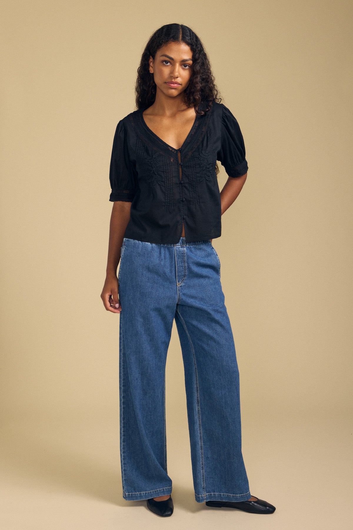 A woman stands against a beige background, wearing the MAVIS TOP by Velvet by Graham & Spencer—a feminine lace blouse with short sleeves—paired with wide-leg blue jeans and black flats, her expression neutral.-41802937991361