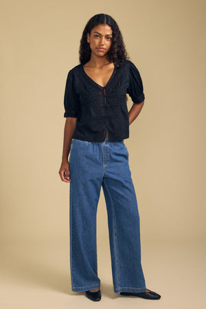 A woman stands against a beige background, wearing the MAVIS TOP by Velvet by Graham & Spencer—a feminine lace blouse with short sleeves—paired with wide-leg blue jeans and black flats, her expression neutral.