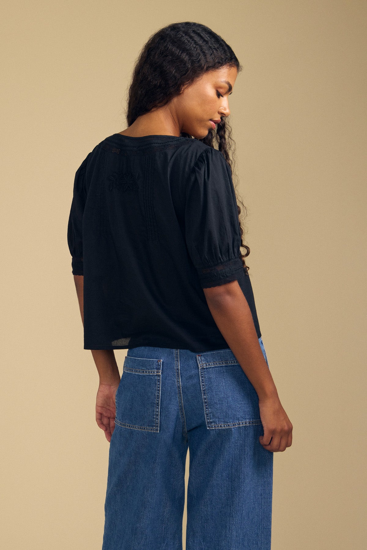 A woman with long curly hair stands facing away, wearing the MAVIS TOP—a feminine black lace blouse by Velvet by Graham & Spencer—paired with blue jeans featuring large back pockets, all set against a plain beige background.-41802937958593