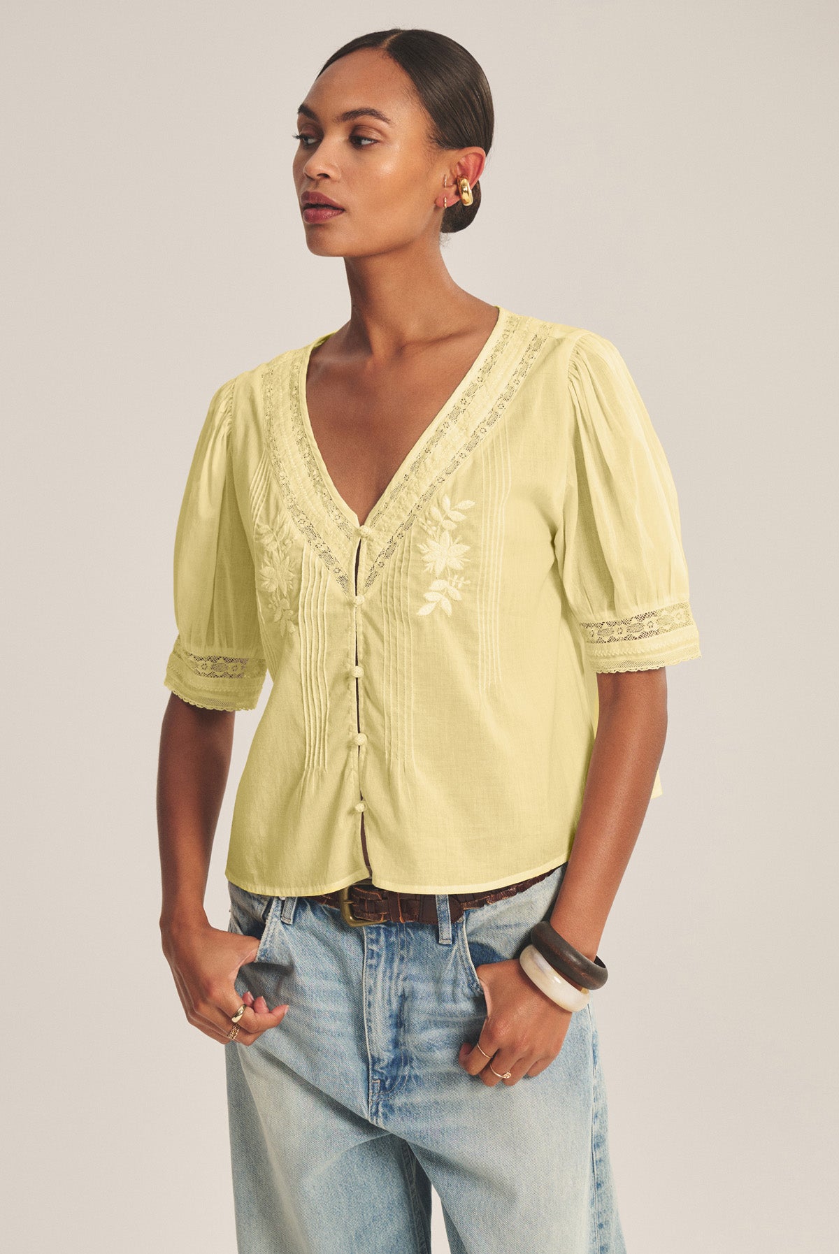 A woman wears the MAVIS TOP by Velvet by Graham & Spencer—a feminine lace blouse with light yellow embroidery and short puffed sleeves—paired with blue jeans, standing with hands in pockets against a neutral background.
