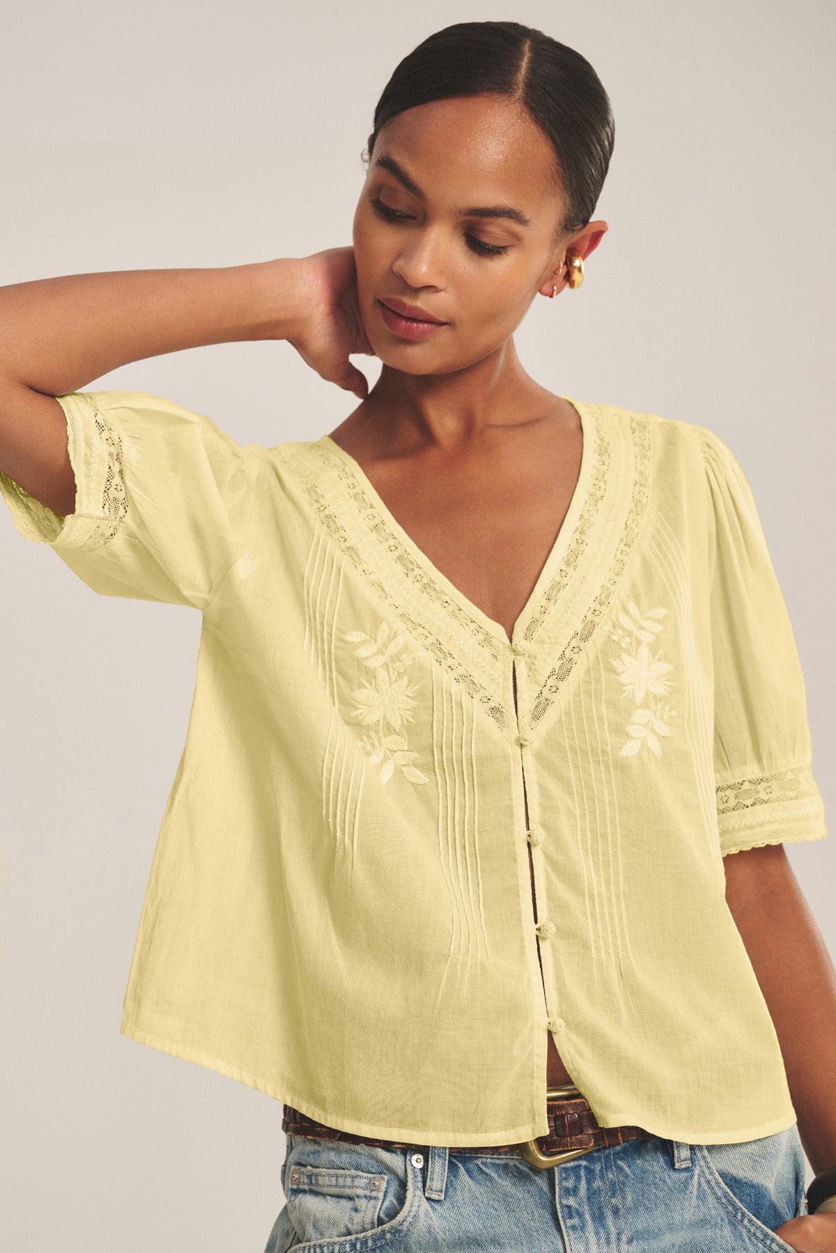 A woman wears the MAVIS TOP by Velvet by Graham & Spencer—a light yellow lace blouse with embroidery—paired with blue jeans, posing with one hand behind her head.