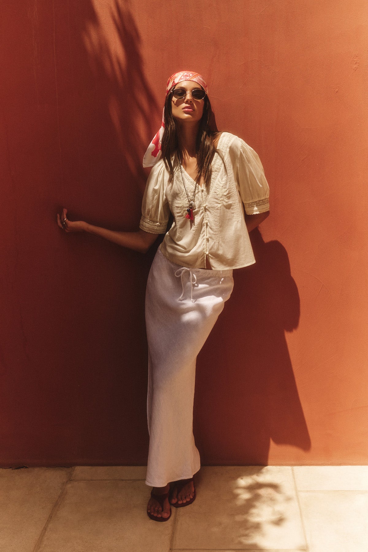 A woman in sunglasses, a pink headscarf, a white short-sleeve blouse, and the BEKA HEAVY LINEN SKIRT by Velvet by Graham & Spencer stands against a terracotta wall in sunlight.