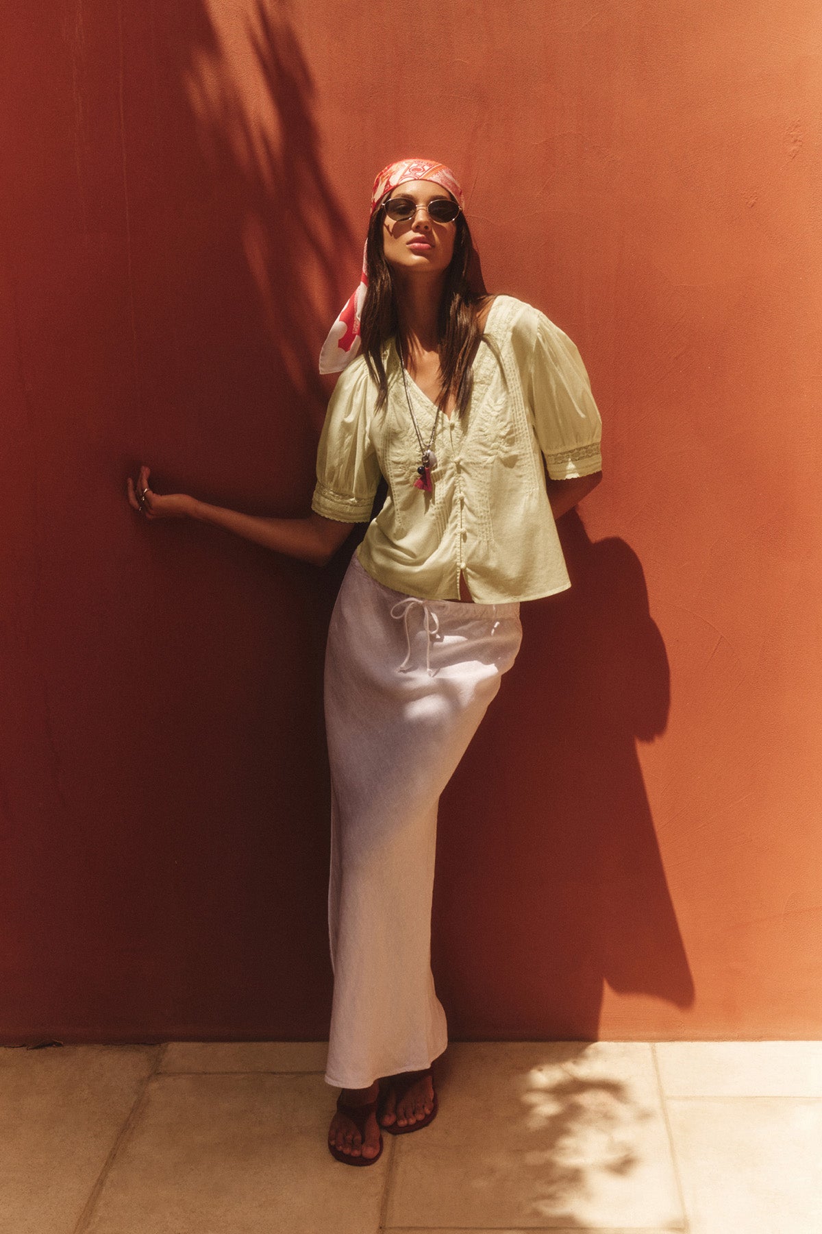 A woman poses casually against a terracotta wall, wearing sunglasses, a red headscarf, white skirt, sandals, and the MAVIS TOP by Velvet by Graham & Spencer. One arm is outstretched in her relaxed stance.