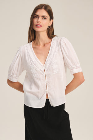 A woman wears the MAVIS TOP by Velvet by Graham & Spencer, a white, short-sleeve cotton lace top with delicate embroidery, paired with a black skirt against a plain background.