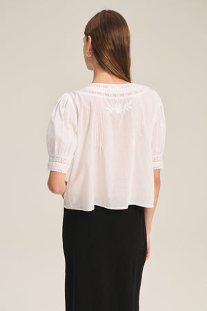 A woman with long brown hair models the MAVIS TOP by Velvet by Graham & Spencer, a feminine lace shirt with back embroidery, paired with a black skirt against a plain background.