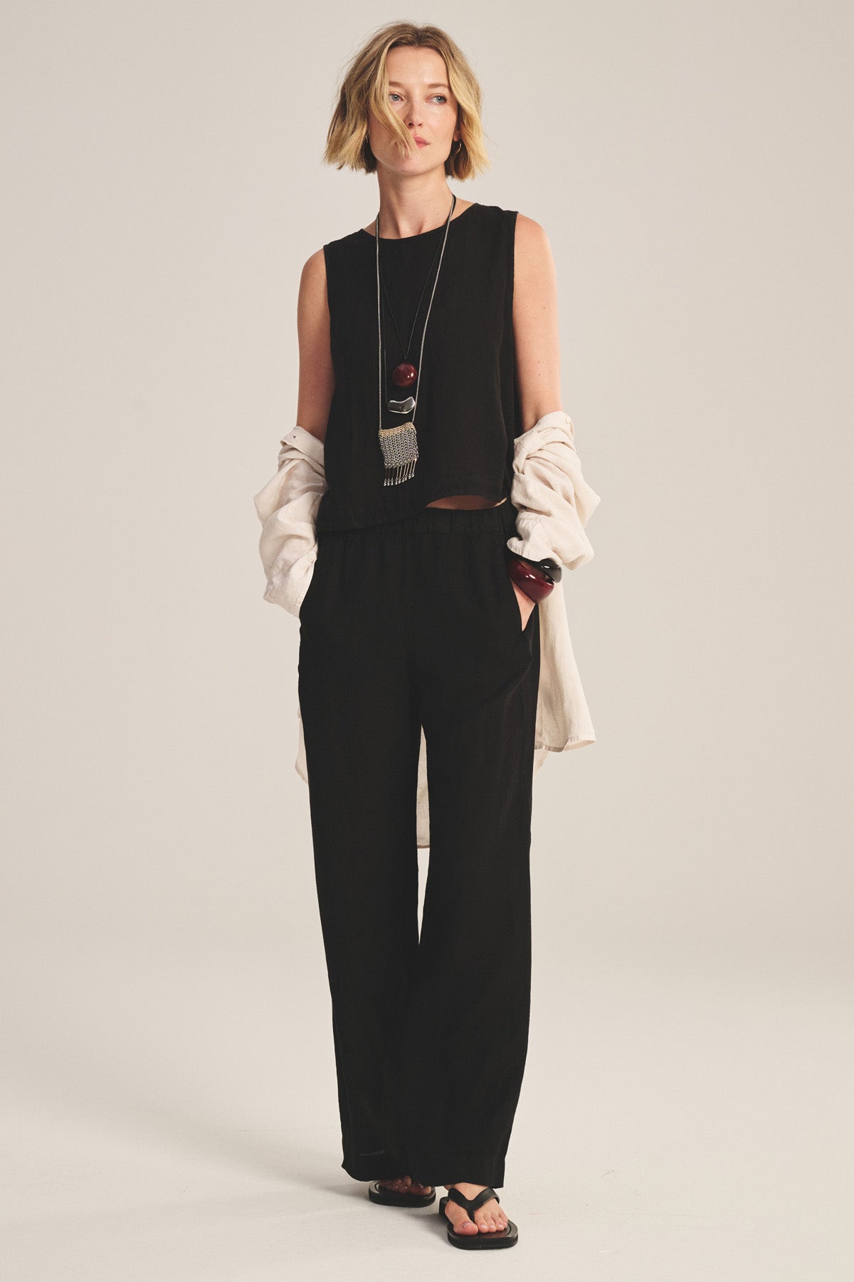 A person wears the MAXINE LINEN TOP by Velvet by Graham & Spencer, paired with black wide-leg pants, a cream jacket draped over their arms, a long necklace, and black sandals.-40808421392577