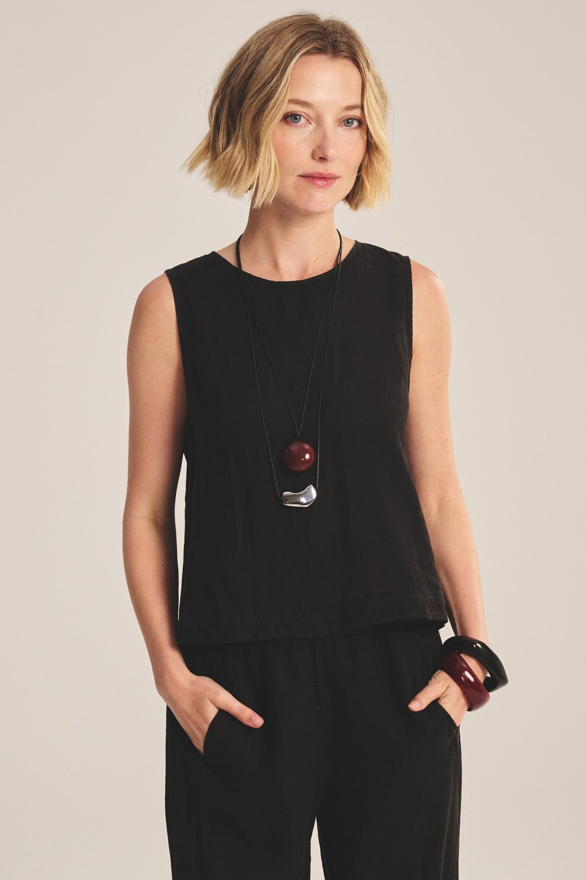 A woman with short blonde hair models the MAXINE LINEN TOP by Velvet by Graham & Spencer, styled sleeveless with black pants and bold jewelry, standing hands-in-pockets against a plain background.