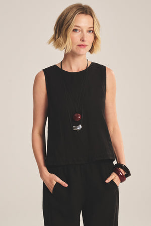 A woman with short blonde hair models the MAXINE LINEN TOP by Velvet by Graham & Spencer, styled sleeveless with black pants and bold jewelry, standing hands-in-pockets against a plain background.