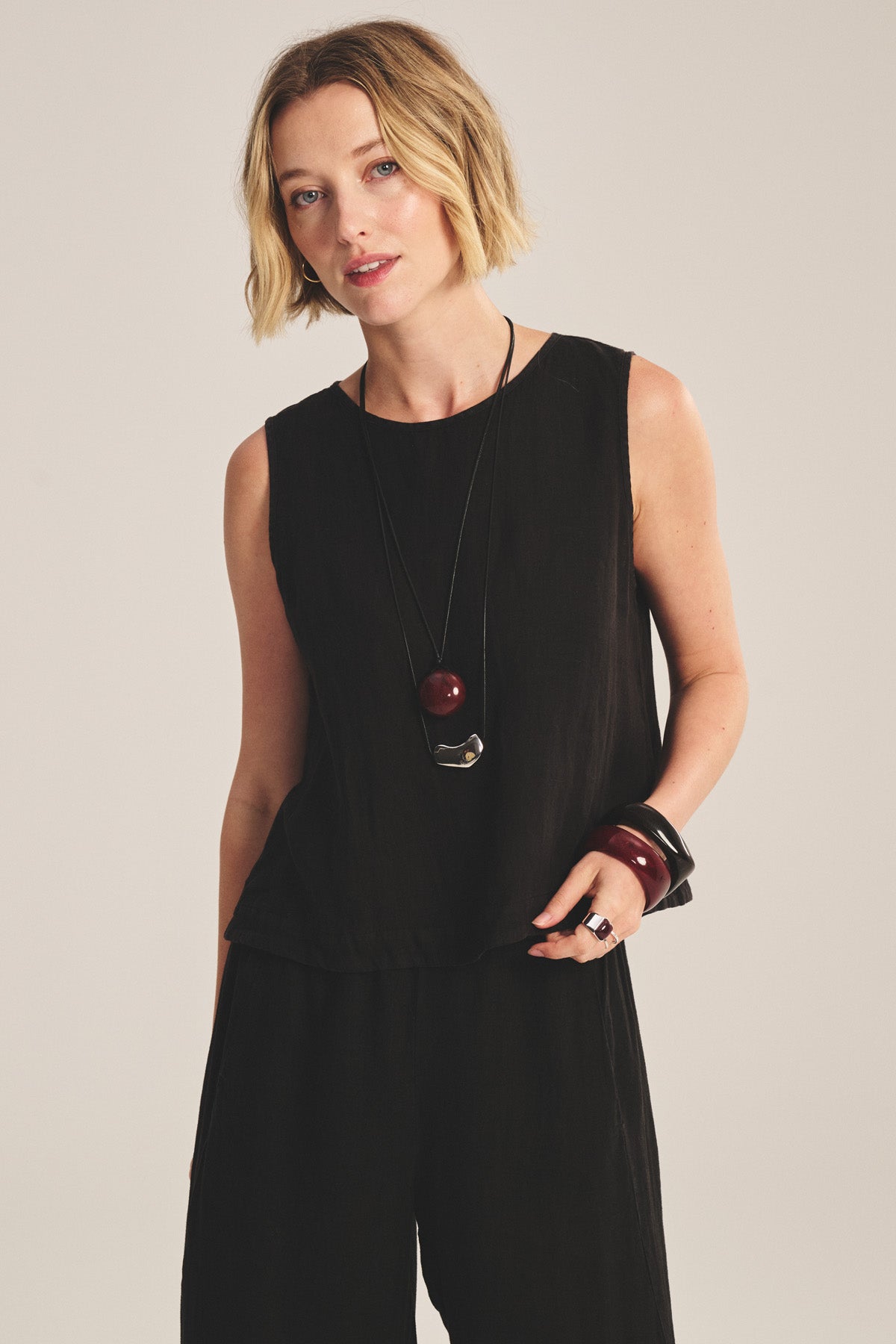 A woman with short blonde hair wears the MAXINE LINEN TOP by Velvet by Graham & Spencer, paired with layered bead necklaces and chunky bracelets, as she poses against a plain background.
