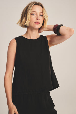 A woman with short blonde hair wears the MAXINE LINEN TOP by Velvet by Graham & Spencer—a sleeveless black linen piece—standing with one hand behind her neck, a neutral expression, and two wide bracelets on her left wrist.
