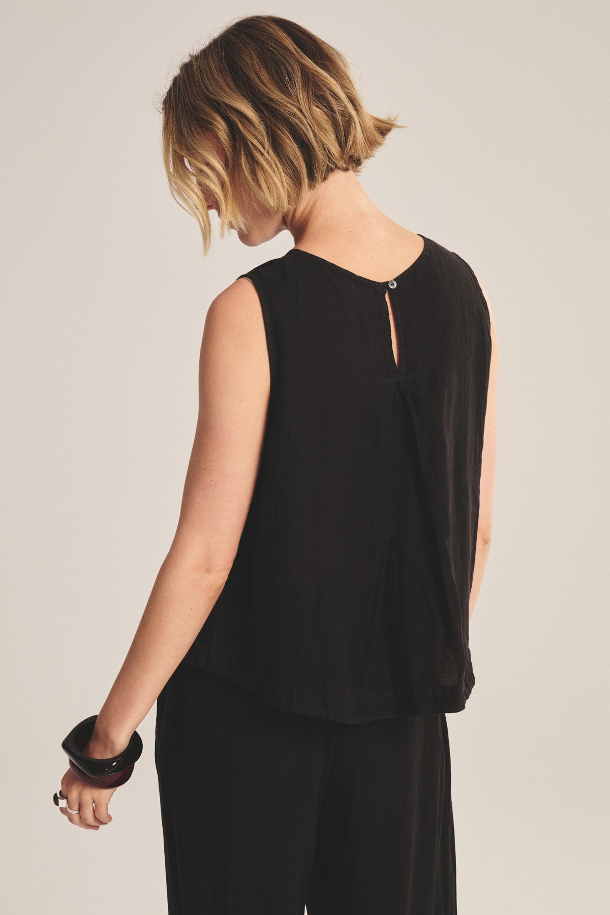 A person with short, wavy hair is seen from behind wearing the MAXINE LINEN TOP by Velvet by Graham & Spencer and black pants, holding a dark bangle in their left hand against a plain background.