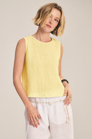 A woman wearing the MAXINE LINEN TOP by Velvet by Graham & Spencer, a sleeveless yellow blouse, paired with white pants and a chain belt, stands against a plain background.