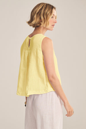 A woman with short blonde hair models the MAXINE LINEN TOP by Velvet by Graham & Spencer—a loose, sleeveless linen blouse—paired with light pants, standing in a side pose against a neutral background.