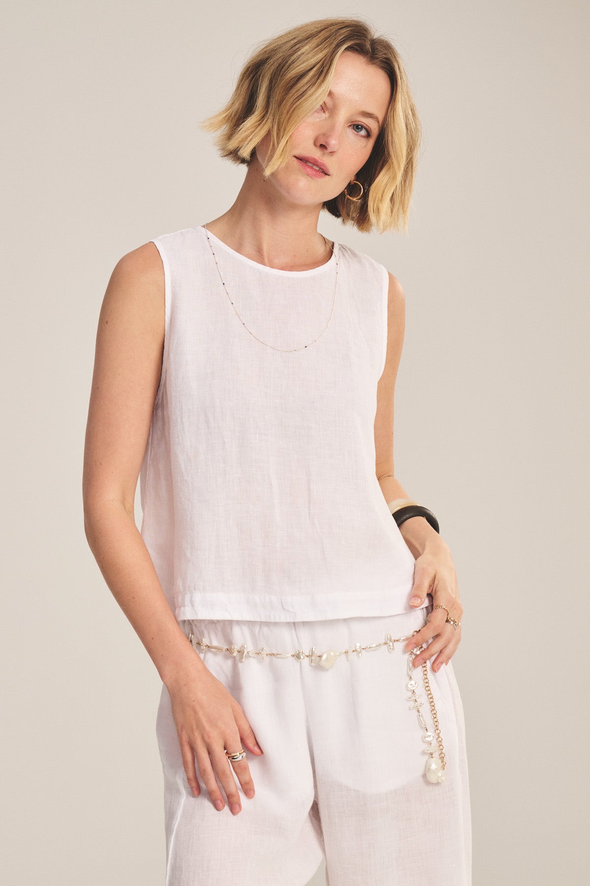 A woman with short blonde hair wears the MAXINE LINEN TOP by Velvet by Graham & Spencer and matching pants, accessorized with a delicate chain necklace and a pearl belt.-41089021313217