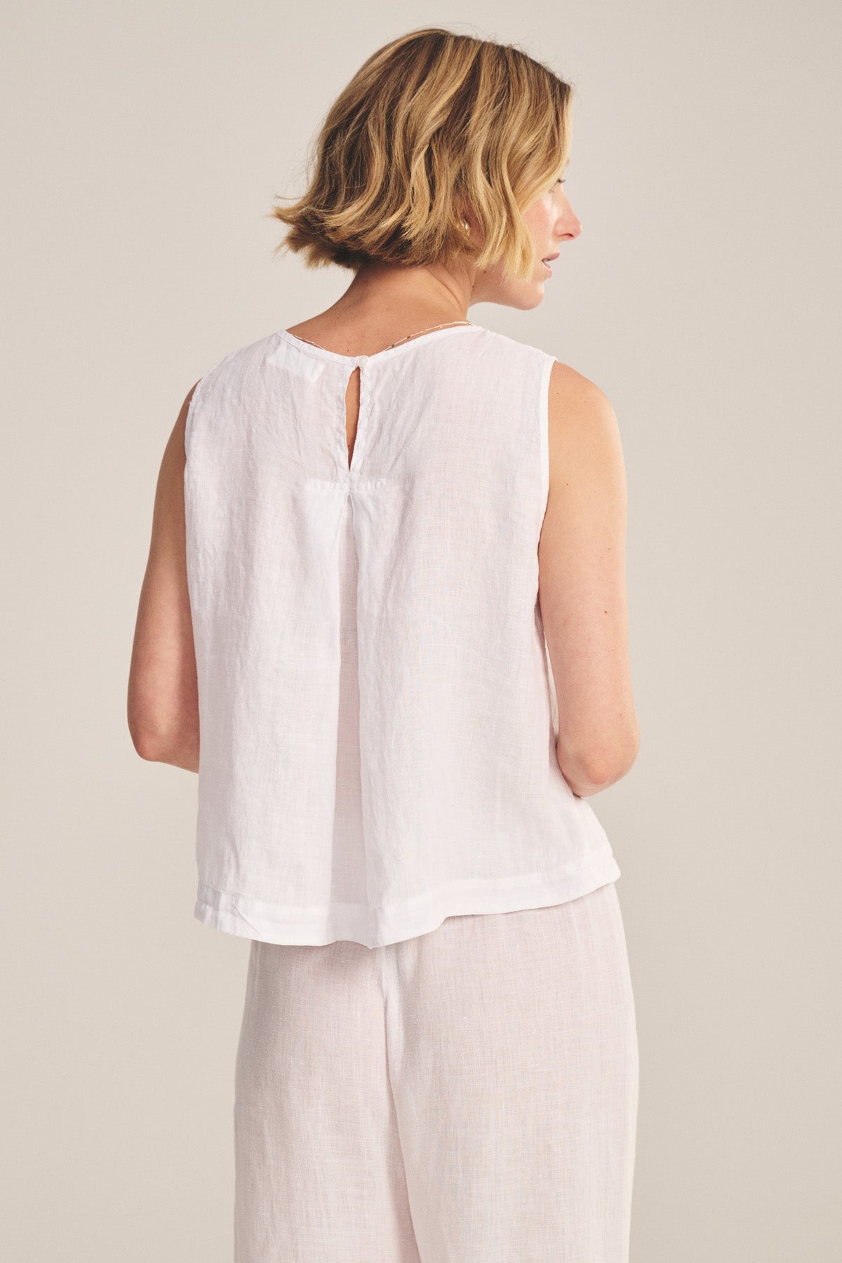 A woman with short blonde hair is seen from behind, wearing the MAXINE LINEN TOP by Velvet by Graham & Spencer and matching loose white pants against a neutral background.